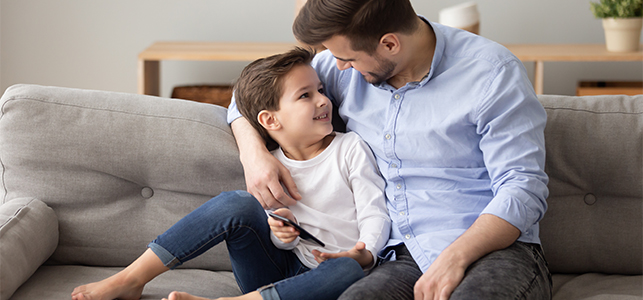 Happy young dad relax sit on couch with preschooler son holding smartphone have fun playing game together, smiling loving father and little boy kid rest on sofa at home spend time using cellphone